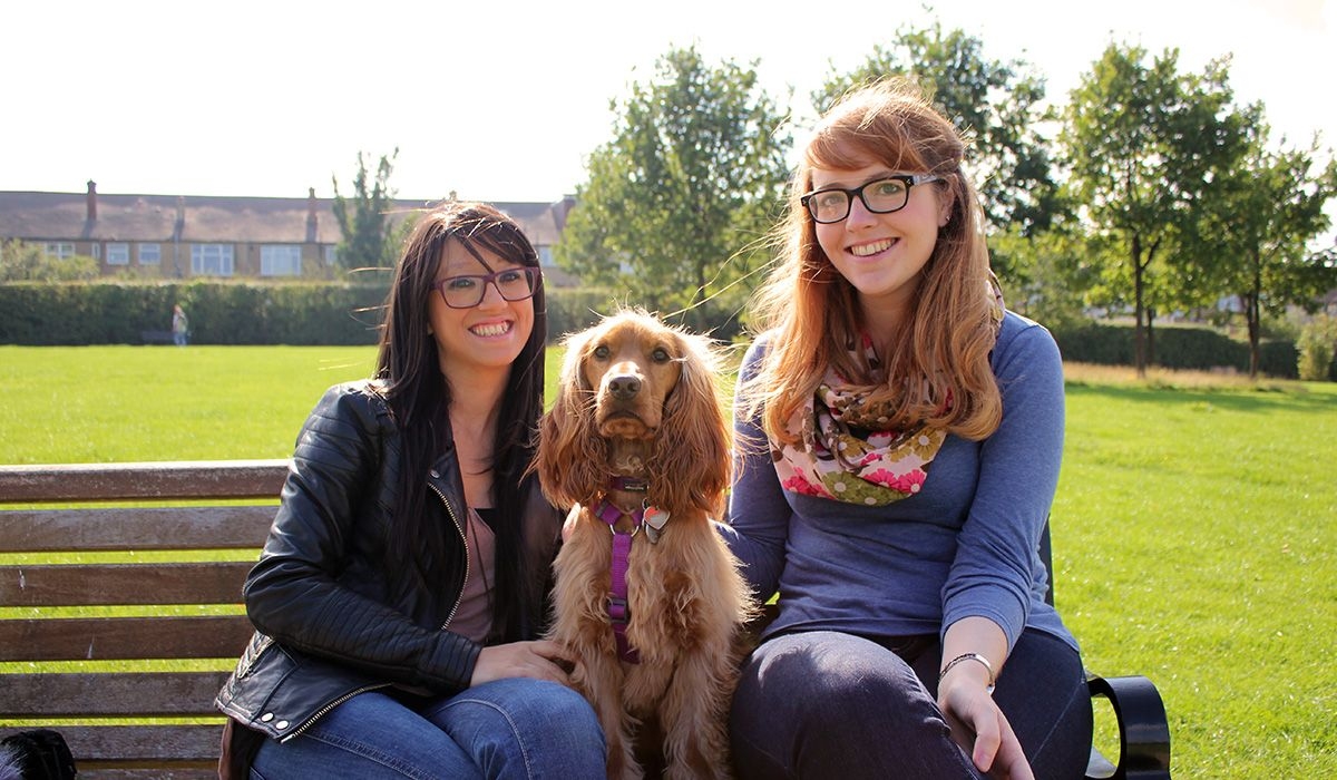 Two women are sitting on a bench in a park on a sunny day, between them is a blonde, long-haired dog