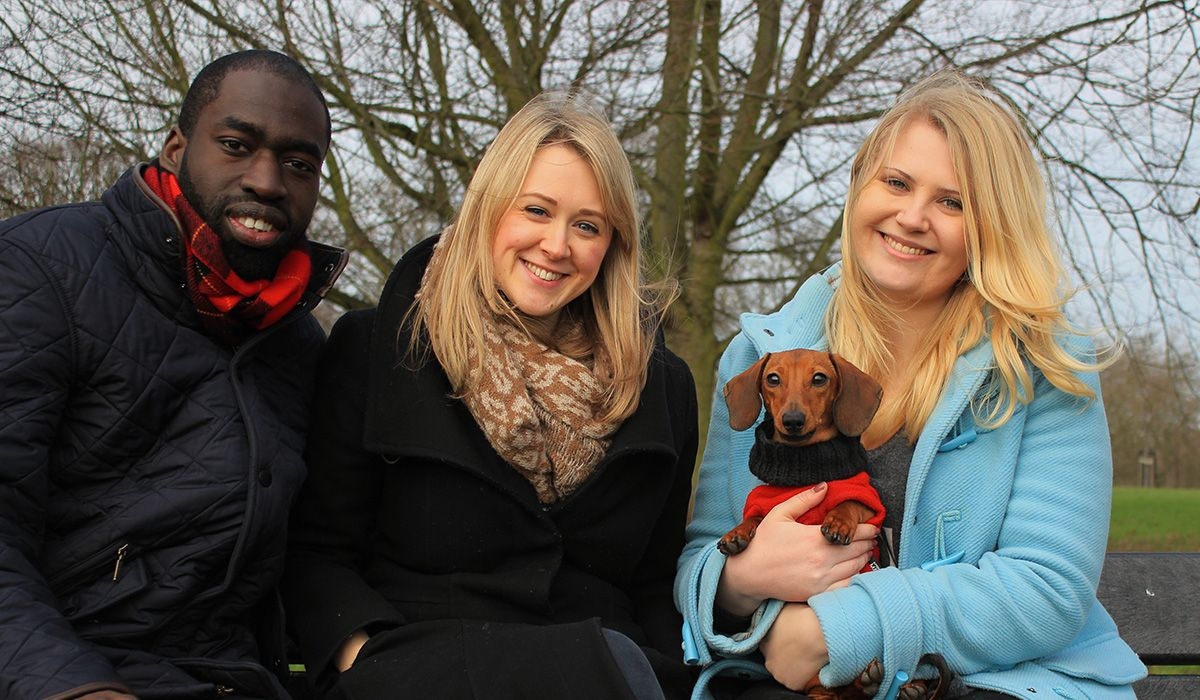 Three people are standing in a park. One of them is holding a small, short-haired, tan dog with large floppy ears