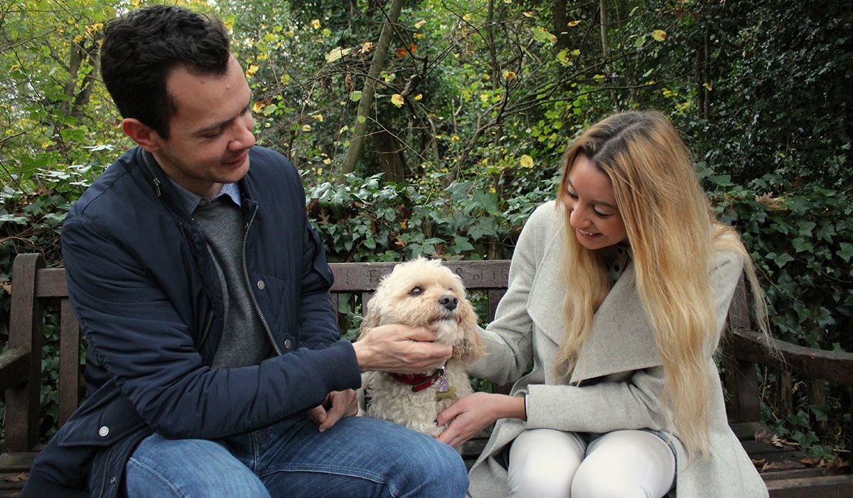 Two people are sitting in a wood on a bench giving loads of attention to a white doggy