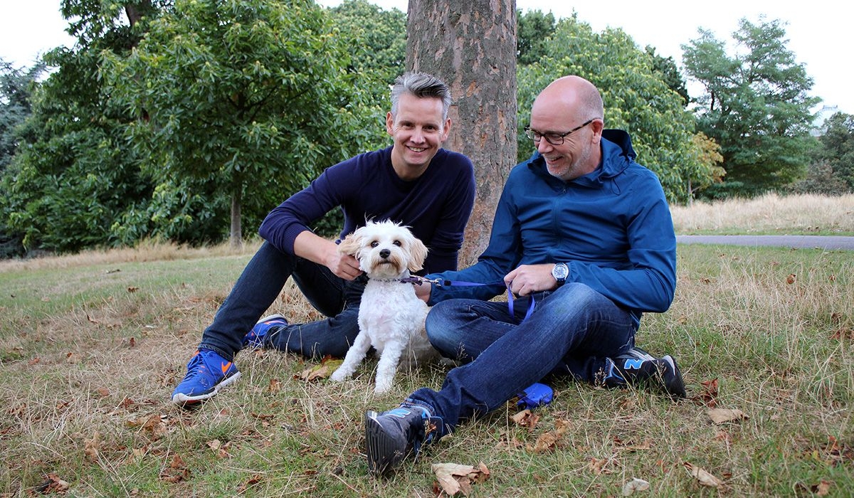 Two men and a small, white dog are sitting on grass under a tree in a park