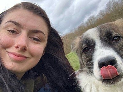 A smiling woman and a dog licking its own nose are posing for a selfie out on a walk