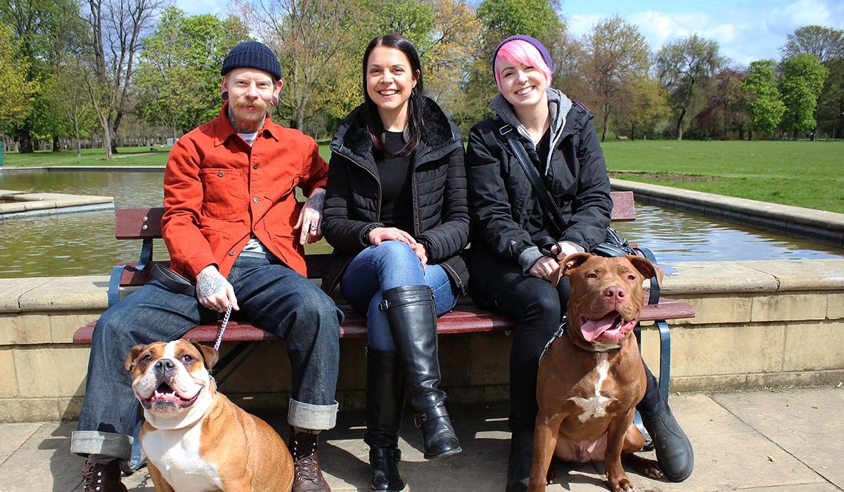 Three smiling people and two dogs are sitting in a park in front of a water feature