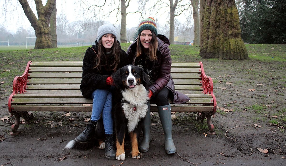 Two women sit on a park bench, smiling. Sitting on the ground between them is a large dog with a thick white, black and tan coat.