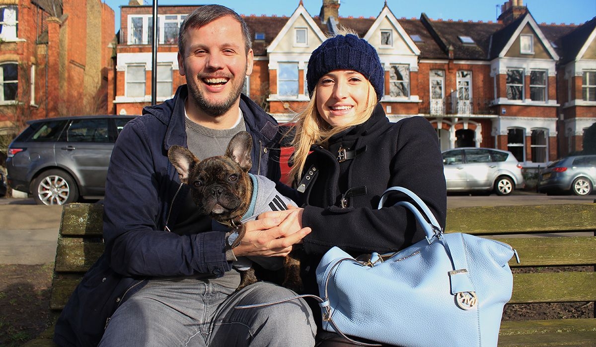 A small dog with a short nose and huge ears is sitting on the laps of two people on a bench in a residential area