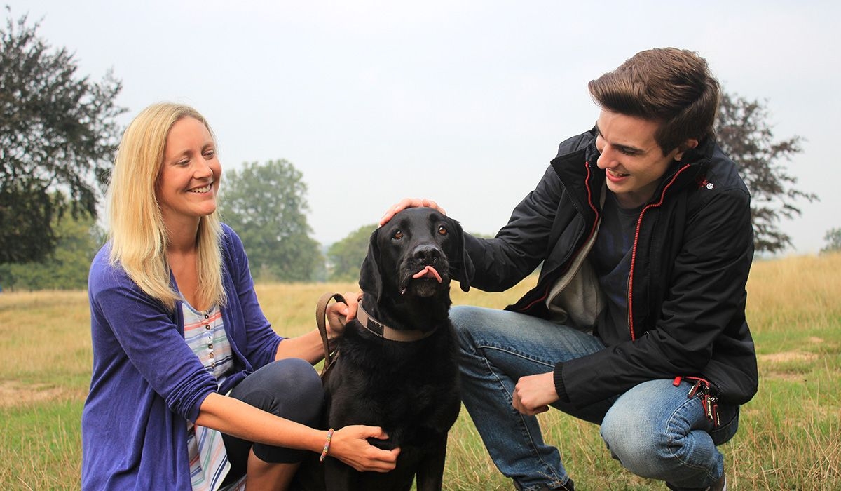 Oso, the black Lab is flanked by two people who are paying attention to him. They are all in the park and all look happy.