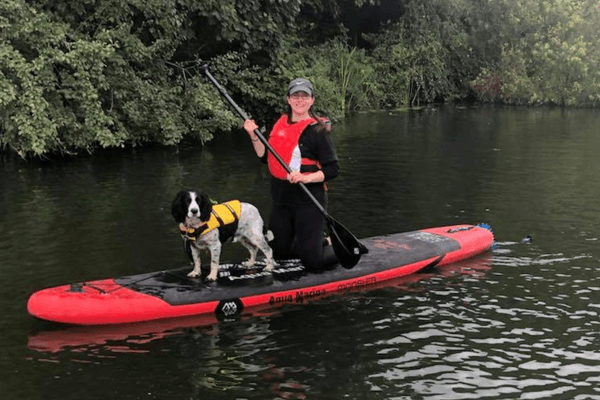 Nicola is kneeling on a red paddleboard, wearing a matching life jacket, with Sprocker, Bob.