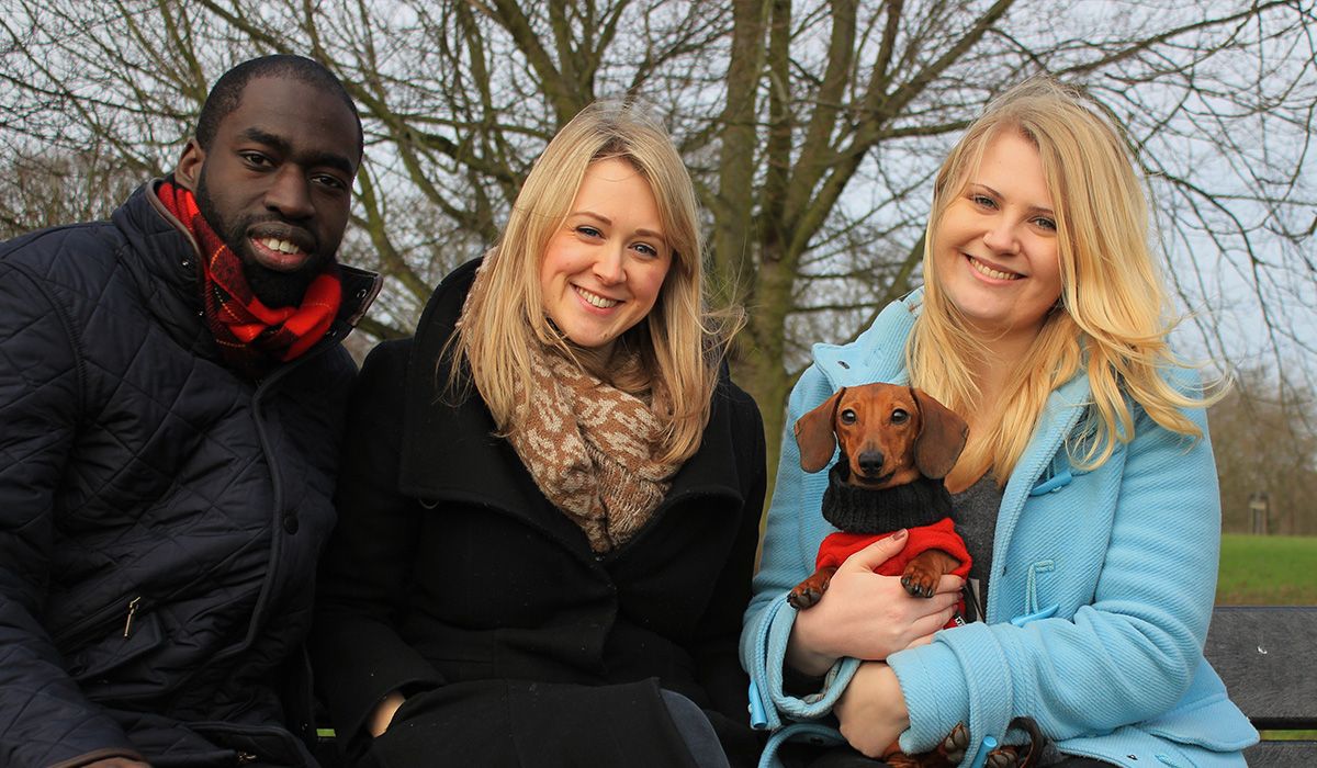 Three people in a park. One of them is holding a small, short-haired, tan dog